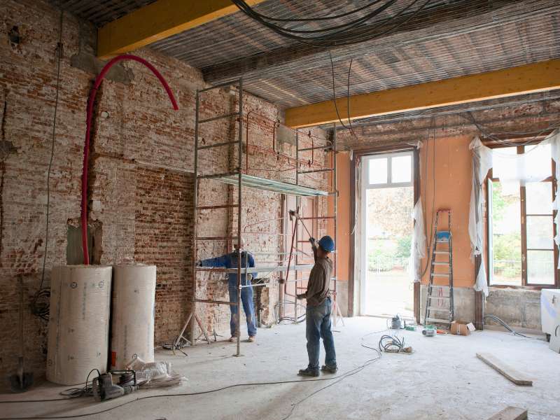 Baustellenraum mit zwei Bauarbeitern, Gerüst und Baumaterialien im Arbeitsbereich.