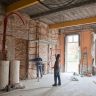 Baustellenraum mit zwei Bauarbeitern, Gerüst und Baumaterialien im Arbeitsbereich.